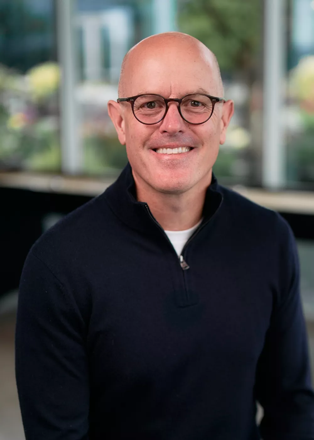 A man wearing a navy blue sweater over a white shirt is seated indoors. The setting features large windows with a blurred view of greenery and modern architecture in the background. The lighting is soft and natural, creating a professional and relaxed atmosphere.